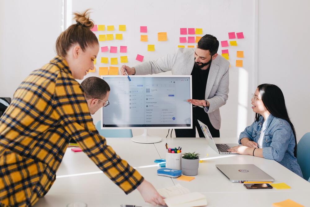 Jeunes gens qui font une réunion autour d'un bureau, en fond des post-it, l'un d'eux montre un ordinateur